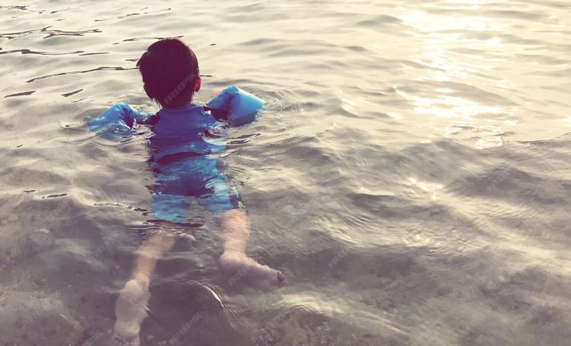 Menino de costas, usando colete ou boias de braço azuis, flutuando em águas rasas do mar na praia. A imagem remete a dicas de segurança para evitar afogamento de crianças.