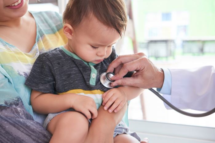 Um médico examinando o coração de um bebê com um estetoscópio. A mãe do bebê está visível, transmitindo a importância do acompanhamento e da prevenção de doenças cardíacas na infância.