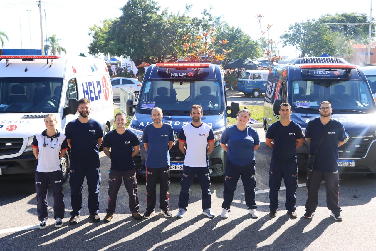 Uma equipe de profissionais da Help Emergências Médicas posa sorrindo em frente a ambulâncias da empresa. A imagem representa o serviço de atendimento e a dedicação da equipe com o cuidado de idosos e toda a família.